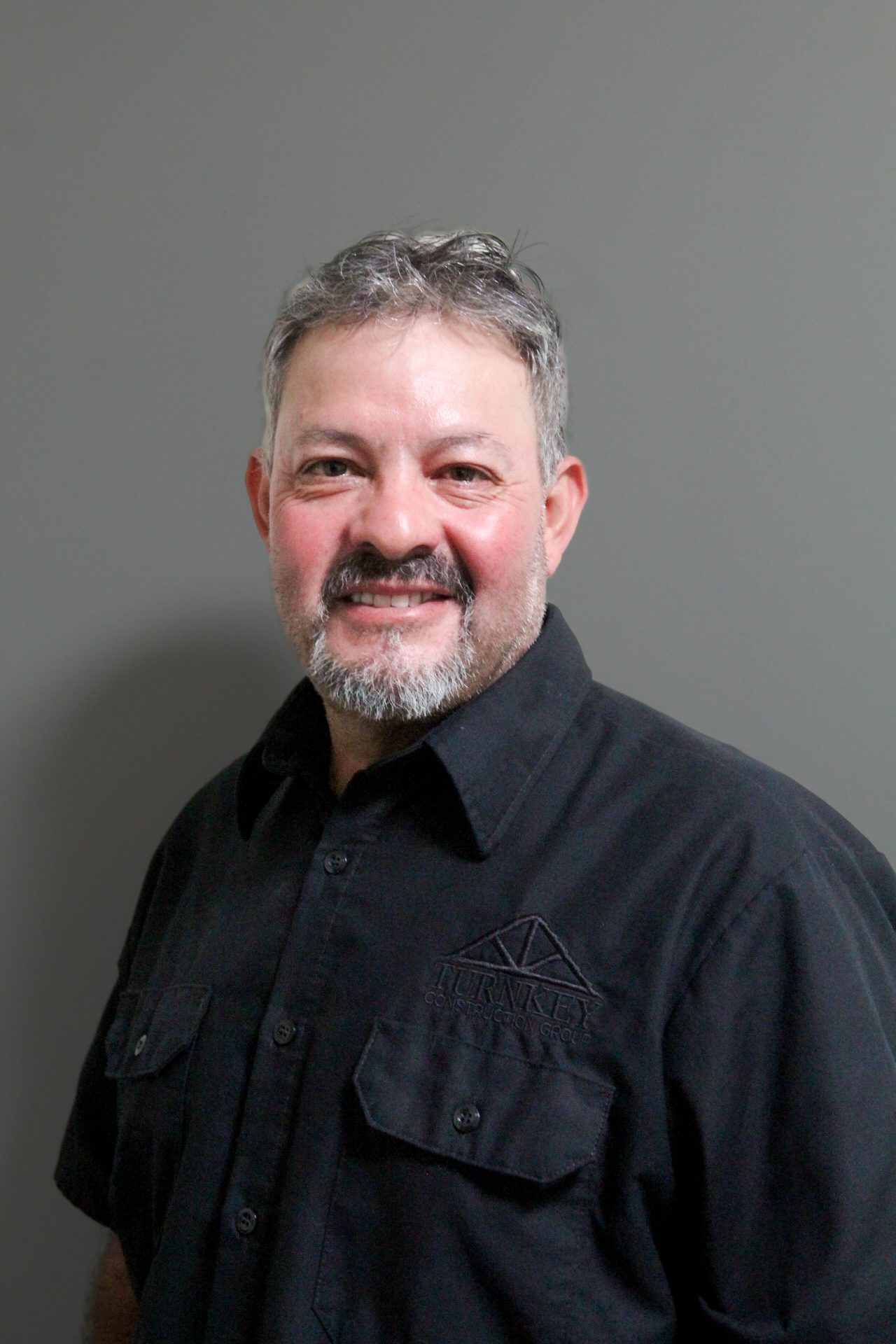 A man in a black shirt smiling in front of a gray wall.